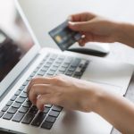 Hands typing on a laptop keyboard while holding a credit card above the trackpad, suggesting online shopping